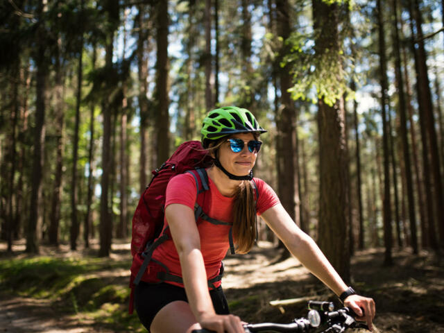 Mountainbikerin mit rotem Shirt, Sonnenbrille und grünem Helm fährt durch den Wald