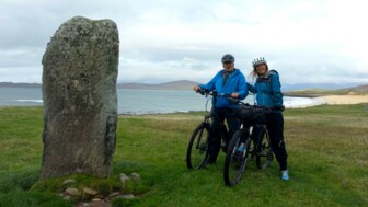Zwei Fahrradfahrer*innen unterwegs auf dem Hebridenweg: Auf einer Wiese stehend mit dem Meer im Hintergrund.