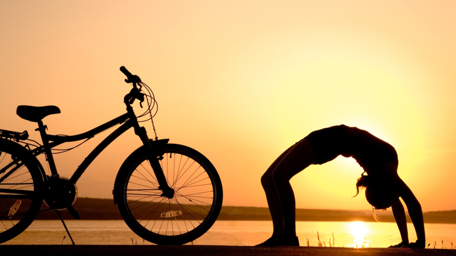 Eine Mountainbikerin in einer Yogaposition in einer Brückenhaltung mit dem Mountainbike nebenan bei Sonnenuntergang an einem See.