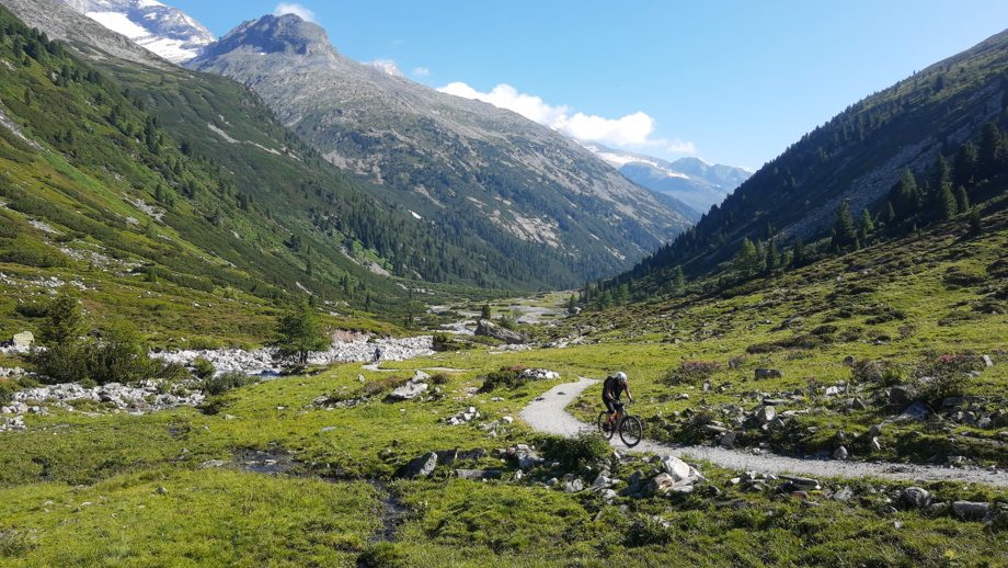 Ein Mountainbiker unterwegs auf der Transalp vom Tegernsee zum Molvenosee mit der Bergen im Hintergrund entlang eines Bachlaufes fahrend.