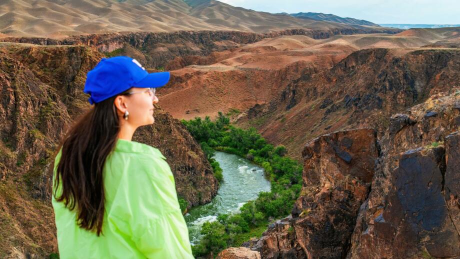 Frau mit grtüner Jacke und blauem Käpp an einem Aussichtspunkt über dem Charyn Canyon mit Fluss und Buschwerk an seinen Ufern