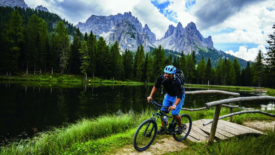 Ein Mountainbiker überquert eine Brücke mit den Dolomiten bei Bewölkung im Hintergrund.