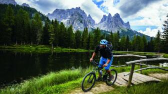 Ein Mountainbiker überquert eine Brücke mit den Dolomiten bei Bewölkung im Hintergrund.