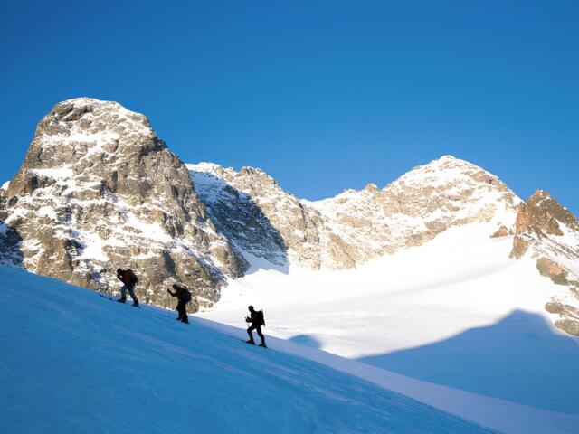 Drei Bergsteiger steigen in gleichmäßigem Abstand eine verschneite, sonnenbeschienene Flanke im Hochgebirge hinauf.