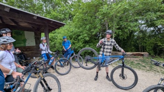 Ein Mountainbike Guide steht in der Mitte der Mountainbikegruppe und zeigt eine neue Übung für das MTB-Fahrtechniktraining vor. Die Gruppe ist im Wald unterwegs.