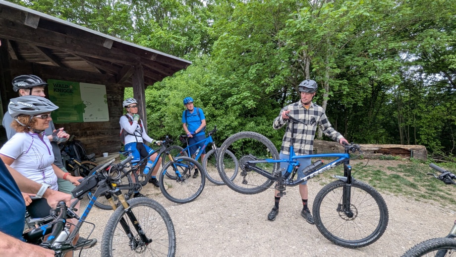 Eine MTB-Gruppe stehend im Wald an einer Hütte. Der Bike -Guide steht in der Mitte der Gruppe und zeigt eine Übung für das MTB-Fahrtechniktraining an.