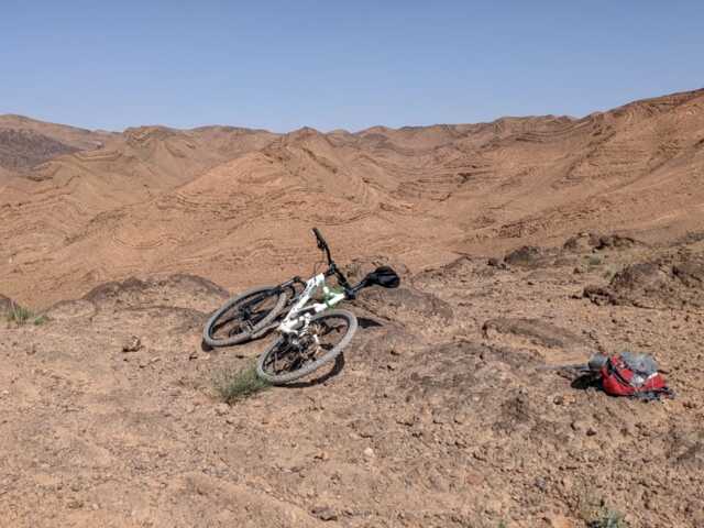 Ein Mountainbike liegt am Wegesrand in der Wüste. Die Wüstenvegetation ist im Hintergrund zu sehen. Die Sonne scheint.