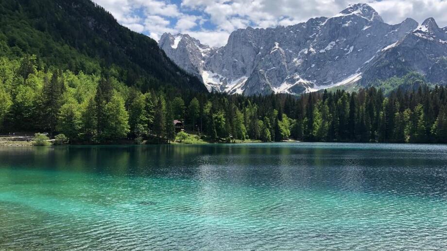 Ein türkisklarer See in Slowenien. Der Himmel ist leicht bewölkt. Die Sonne scheint. Im Hintergrund sind die Berge zu sehen.