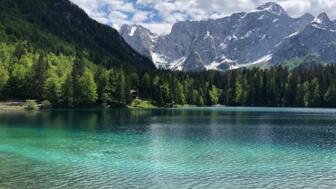 Ein türkisklarer See in Slowenien. Der Himmel ist leicht bewölkt. Die Sonne scheint. Im Hintergrund sind die Berge zu sehen.