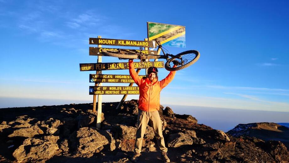 Ein Mountainbiker hebt sein Fahrrad in die Höhe. Er steht am Gipfel des Kilimandscharo. Das "Gipfelkreuz" ist im Hintergrund. Der Himmel ist blau.
