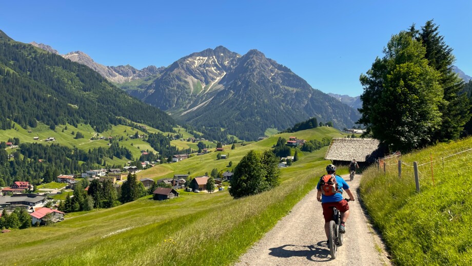 Eine Gruppe an Mountainbiker unterwegs auf Tour.  Sie fahren auf einer Straße. Die Sonne scheint. Im Hintergrund sieht man die Berge.