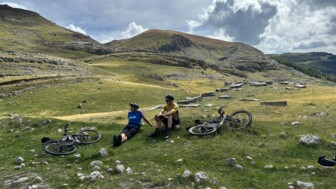 Zwei Mountainbiker machen eine Pause auf einer Wiese. Im Hintergrund sind die Berge.
