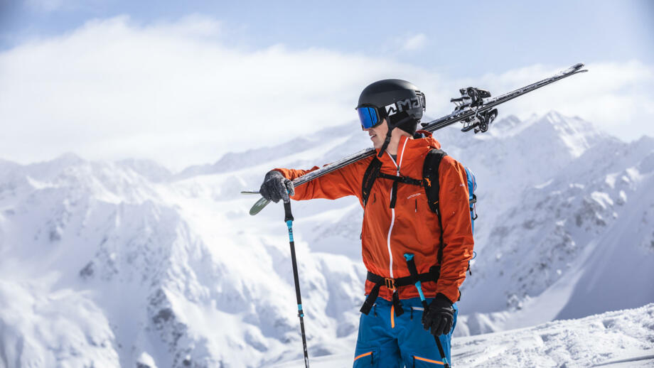 Skifahrer schultert seine Ski, im Hintergrund schneebedeckte Berge