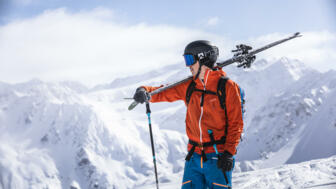 Skifahrer schultert seine Ski, im Hintergrund schneebedeckte Berge