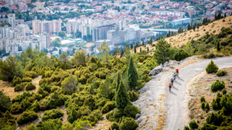 Drei Mountainbiker unterwegs durch Bosnien-Herzegowina. Im Hintergrund sieht man eine Stadt. Die drei Biker radeln auf einer Straße den Berg auswärts.