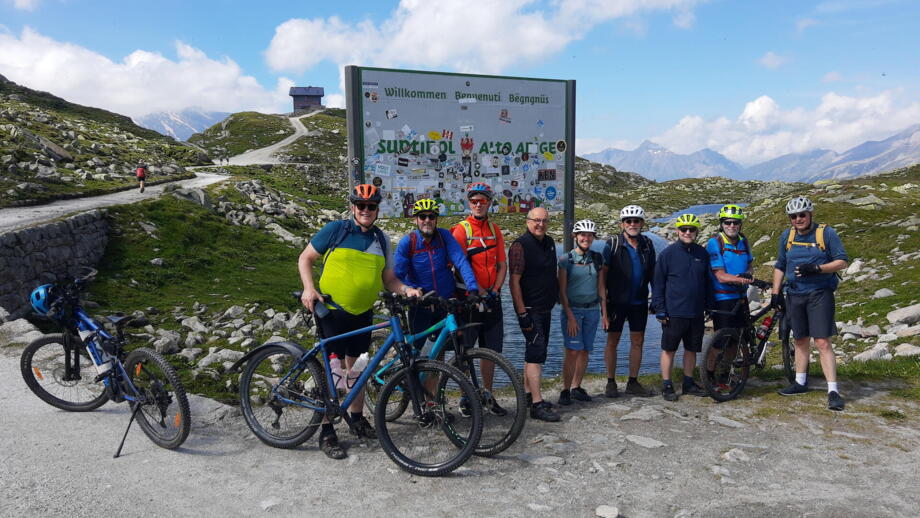 Eine Gruppe von Mountainbiker*innen am Pfitscherjoch. Sie stehen vor dem Schild umliegend ist die Bergwelt.