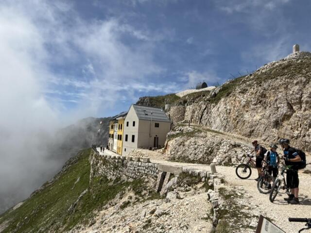 Eine Gruppe an Mountainbiker*innen auf Tour - sie fahren auf einem Pfad Richtung Berghütte. Der Himmel ist bewölkt.
