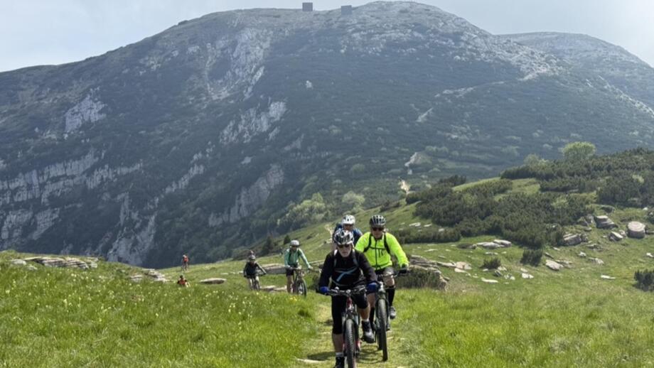 Eine Gruppe an Mountainbiker*innen unterwegs auf der Tour. Sie fahren über eine Bergwiese. Im Hintergrund sind die Berge zu sehen.