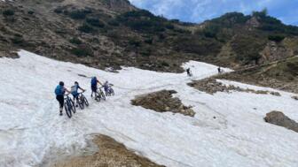 Eine MTB-Gruppe auf Tour. Sie überqueren ein Schneefeld und schieben Ihr MTB über das Schneefeld. Die Sonne scheint. Der Himmel ist bewölkt. Im Hintergrund sind die Berge.