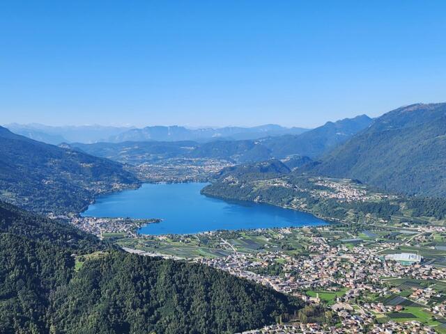 Der Levicosee mit der umliegenden Bergwelt und der Stadt Levico Terme. Die Sonne scheint.