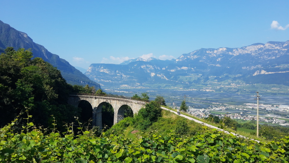 Eine Viaduktbrücken im Hintergrund ist die Bergwelt zu sehen. Die Sonne scheint. Der Himmel ist blau.