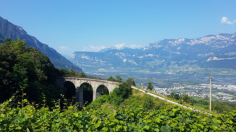 Eine Viaduktbrücken im Hintergrund ist die Bergwelt zu sehen. Die Sonne scheint. Der Himmel ist blau.