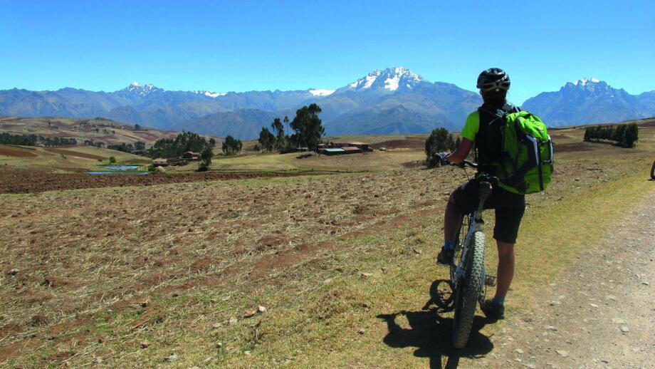 Ein Mountainbiker am Wegesrand stehend. Im Hintergrund sind die Berge, teils schneebedeckt zu sehen. Der Himmel ist blau. Die Sonne scheint.