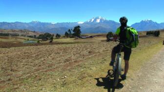 Ein Mountainbiker am Wegesrand stehend. Im Hintergrund sind die Berge, teils schneebedeckt zu sehen. Der Himmel ist blau. Die Sonne scheint.
