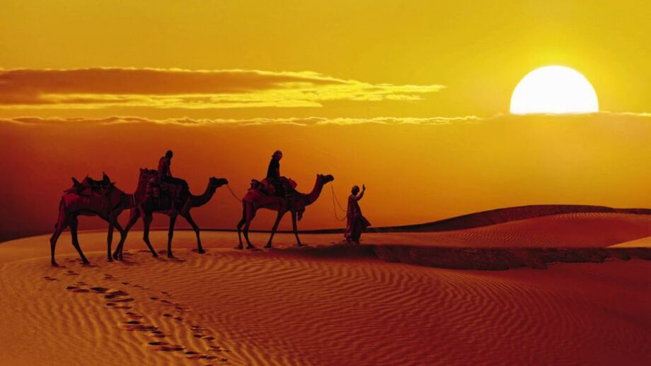 Zwei Personen reiten zum Sonnenuntergang auf Kamelen durch die Wüste Thar und werden von einem Kameltreiber geführt