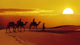 Zwei Personen reiten zum Sonnenuntergang auf Kamelen durch die Wüste Thar und werden von einem Kameltreiber geführt