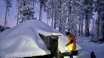 Ein Mann, der in der Dämmerung vor einer schneeverschneiten Hütte steht.