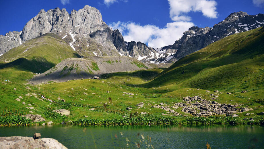 Georgisches Dorf Stepantsminda in grüner Wiesenlandschaft mit See im Vordergrund und felsigen Bergen im Hintergrund