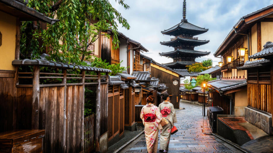 Zwei Frauen in traditionellen Gewändern spazieren durch eine Gasse mit Holz- und Steinhäusern in der Altstadt von Kyoto mit Blick auf eine vierstufige Pagode im Hintergrund