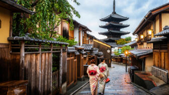 Zwei Frauen in traditionellen Gewändern spazieren durch eine Gasse mit Holz- und Steinhäusern in der Altstadt von Kyoto mit Blick auf eine vierstufige Pagode im Hintergrund