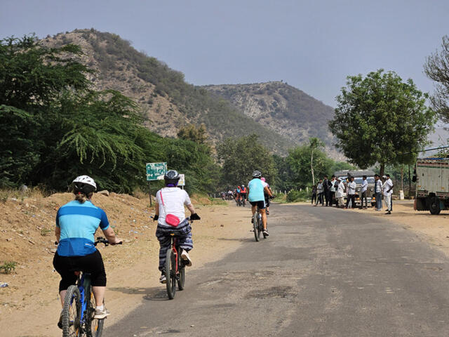 Mehrere Radreisende fahren auf einer Landstraße durch die hügelige Landschaft Rajasthans. Die Strecke führt vorbei an trockenen Berghängen, grünen Bäumen und kleinen Gruppen neugieriger Menschen am Straßenrand. Die Szene vermittelt Abenteuer, Bewegung und das besondere Gefühl einer aktiven Reise durch das ländliche Indien.
