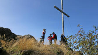 Ein Mountainbike Gruppe an einem Gipfelkreuz. Der Himmel ist blau und die Sonne scheint.