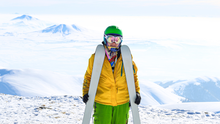 Frau in gelber und roter Skibeschleidung mit Skiern auf dem Taygenis-Gipfel mit Vulkanen im Hintergrund