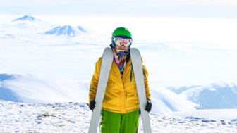 Frau in gelber und roter Skibeschleidung mit Skiern auf dem Taygenis-Gipfel mit Vulkanen im Hintergrund