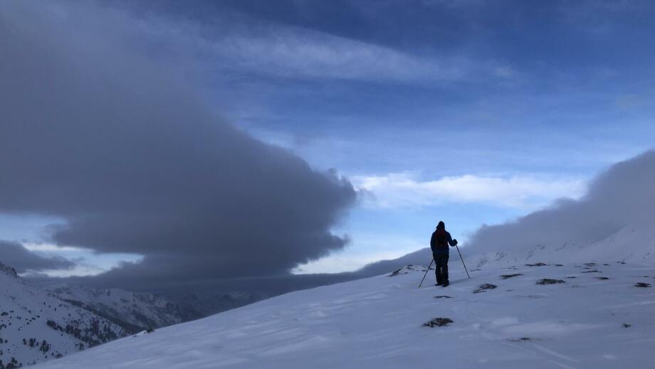 Splitboarder im Aufstieg vor seitlicher Wolkenfront und blauem Himmel.