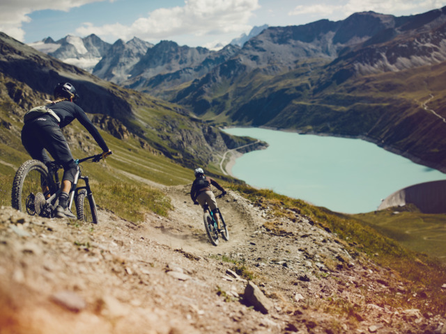 Zwei Mountainbiker*innen während der Abfahrt mit Bergpanorama der Walliser 4000er im Hintergrund.