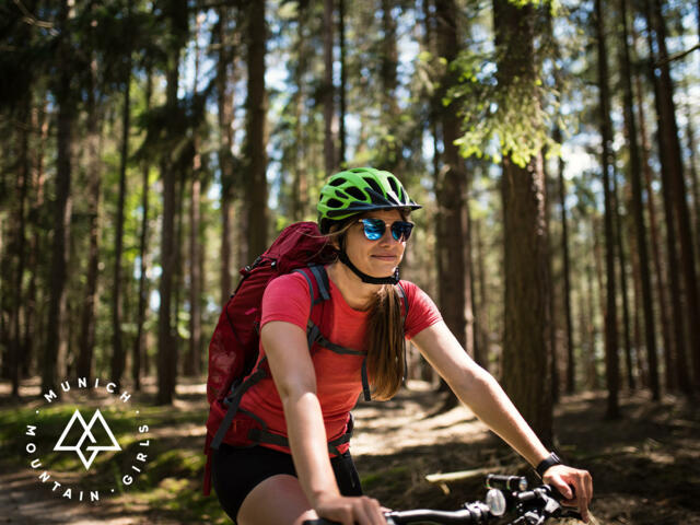 Mountainbikerin mit rotem Shirt, Sonnenbrille und grünem Helm fährt durch den Wald