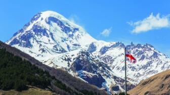 Vergletscherter Fünftausender Kasbek mit grünen und braunen Berghängen im Vordergrund und georgischer Flagge