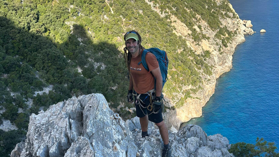 Guide auf der Spitze der Klippe mit Rucksack und Sicherungsgurten und -seilen, im Hintergrund die Küste mit herrlich blauem Meerblick