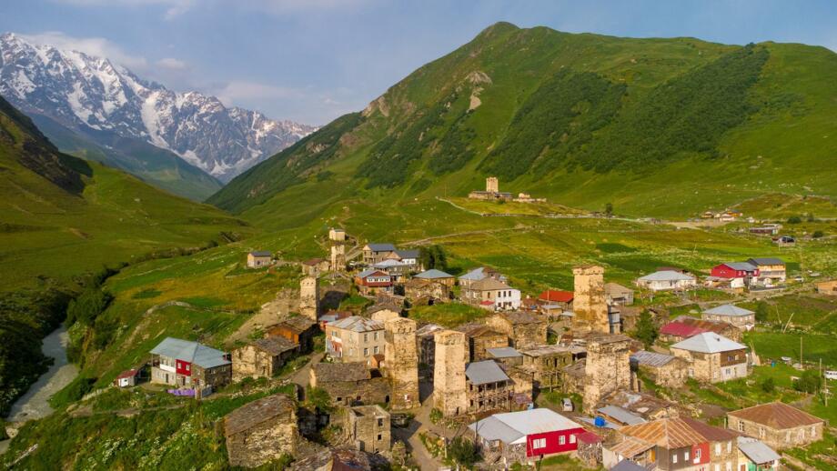 Dorf Ushguli in Swanetien mit Wohnhäusern und Wehrtürmen  in grüner Hügellandschaft mit vergletscherter Kaukasus-Bergkette im Hintergrund