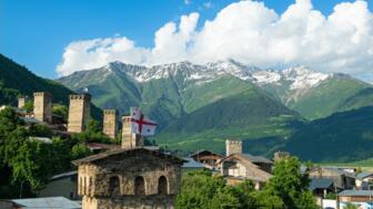 Ort Mestia in Swanetien mit Häusern, Wehrtürmen und einer georgischen Fahne vor grüner und im oberen Bereich verschneiter Bergkulisse