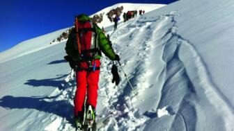 Bergsteigergruppe in teilweiswer farbenfroher Daunenkleidung quert eine Schneeflanke am Fünftausender Kasbek mit Skiern