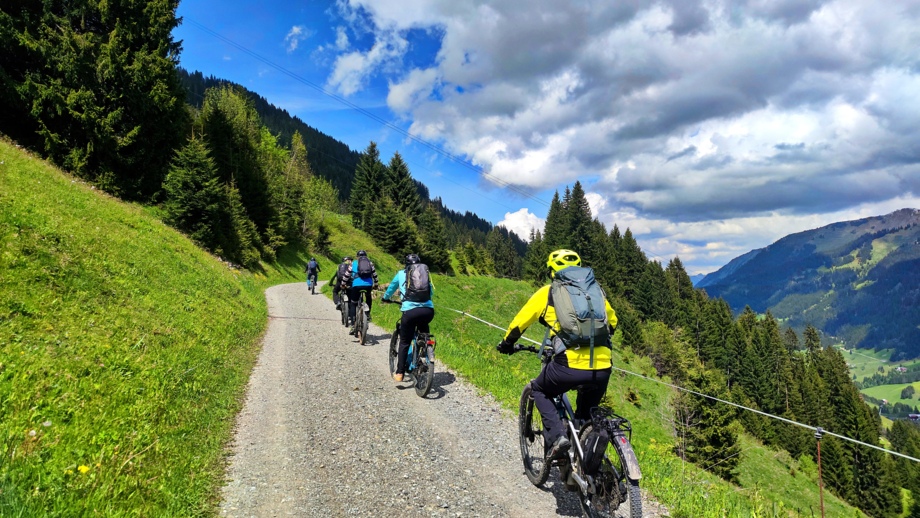Eine Gruppe an E-Mountainbiker unterwegs auf der Tour im Kleinwalser auf einem Höhenweg fahrend. Der Himmel ist bewölkt und die Sonne scheint. Im Hintergrund sieht man die Bergwelt der Allgäuer Alpen.