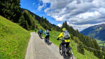 Eine Gruppe an E-Mountainbiker unterwegs auf der Tour im Kleinwalser auf einem Höhenweg fahrend. Der Himmel ist bewölkt und die Sonne scheint. Im Hintergrund sieht man die Bergwelt der Allgäuer Alpen.