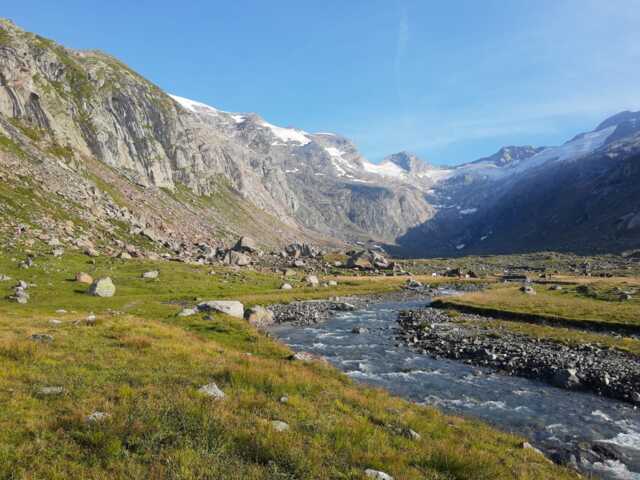 Ein Fluss schlängelt sich durch das Tal. Im Hintergrund ist die Bergwelt zu sehen. Es scheint die Sonne.