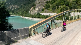 Zwei Mountainbiker überquere einen Fluss über eine Brücke. Im Hintergrund sieht man die Bergwelt.